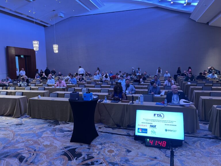Wide-angle view of a traditional conference room with rows of tables, tan linens, and attendees seated under blue-toned lighting.