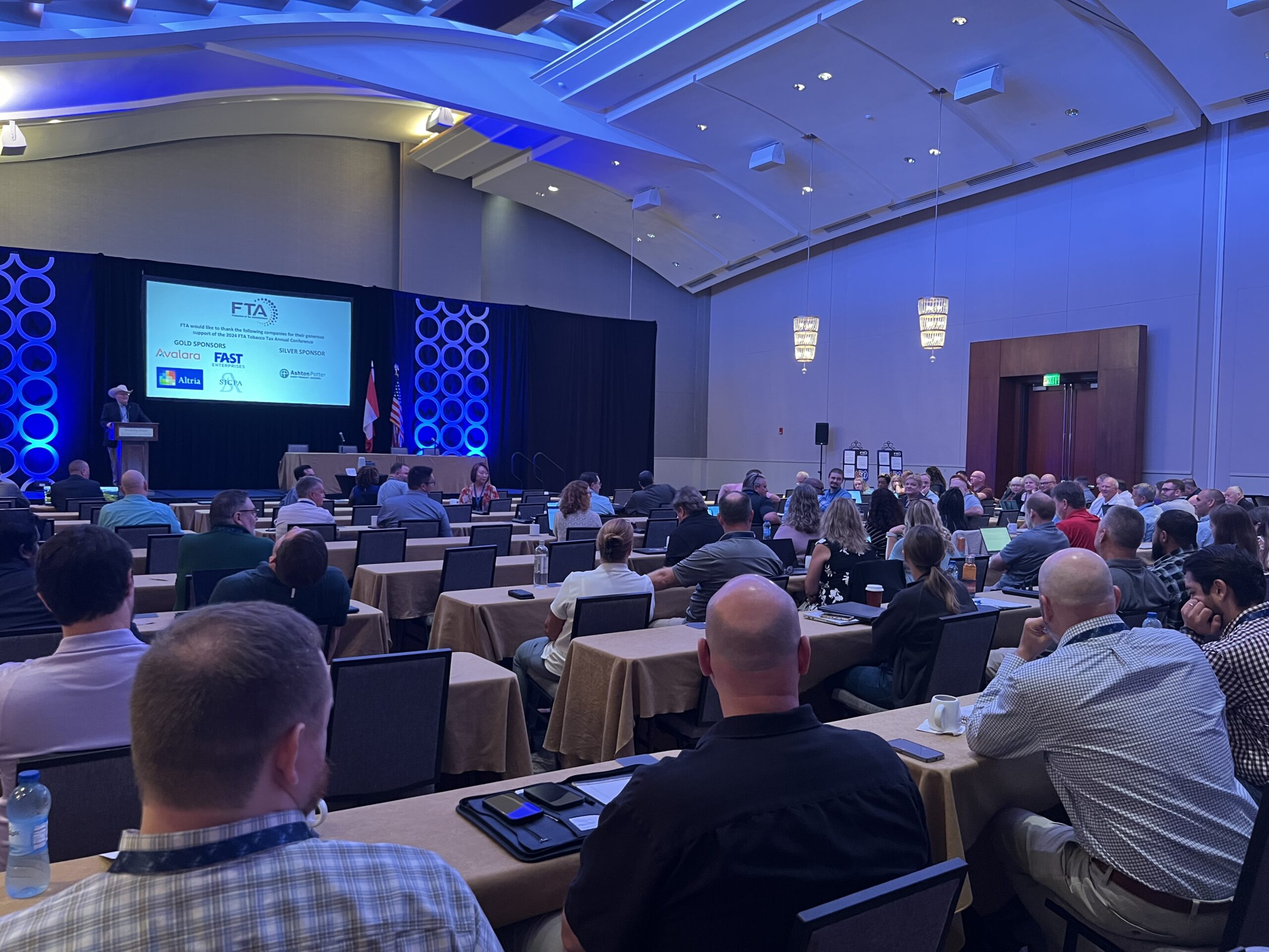 Speaker presenting on stage at a formal conference with projection screen, blue lighting, and rows of attendees watching from tables.