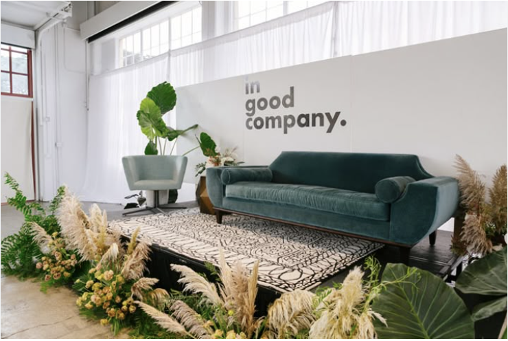 Modern, styled stage for a coaching event with a velvet teal couch, accent chair, lush greenery, and branded ‘In Good Company’ backdrop.